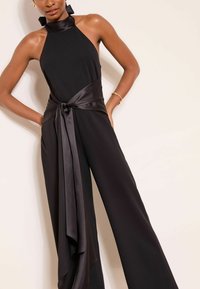 Tuta nera in tessuto liscio con collo all'americana, vita aderente con cinturino in raso e gamba larga per una silhouette fluida.