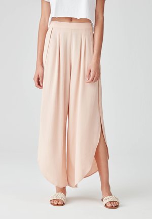 Femme portant un pantalon rose poudré taille haute à jambes larges avec des fentes latérales et des sandales blanches tissées, visibles du nombril jusqu’aux pieds.