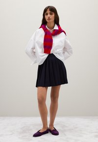 Camisa blanca de gran tamaño, falda azul marino plisada y una bufanda a rayas roja y púrpura. El calzado es plano, casual y de color púrpura oscuro. Fondo simple.