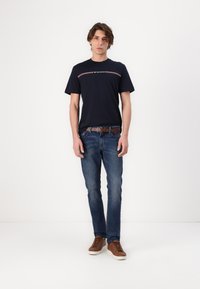 Marineblå kortermet t-skjorte med en rød og hvit horisontal stripe over brystet, kombinert med blå jeans og brune snørebåndssneakers.