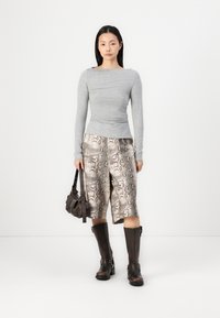 Grijze lange mouw top, slangpatroon shorts, bruine kniehoge laarzen en een kleine bruine handtas. Neutrale achtergrond.