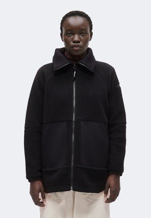 Persona con cabello corto y rizado que lleva una chaqueta de forro polar negra con cremallera y grandes bolsillos frontales, y pantalones de color claro sobre un fondo liso.