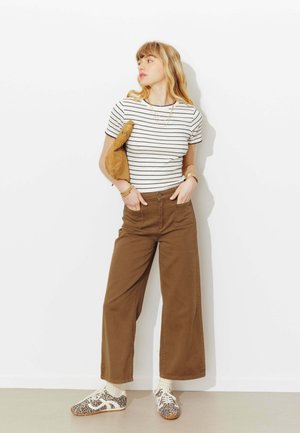 Femme debout contre un mur blanc portant un t-shirt rayé blanc et noir, un pantalon large marron, des baskets imprimé léopard, tenant un sac en daim beige.