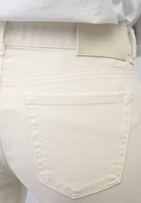 Jeans de mezclilla color crema con un bolsillo trasero y una etiqueta de marca de cuero con textura y en relieve en la cintura.
