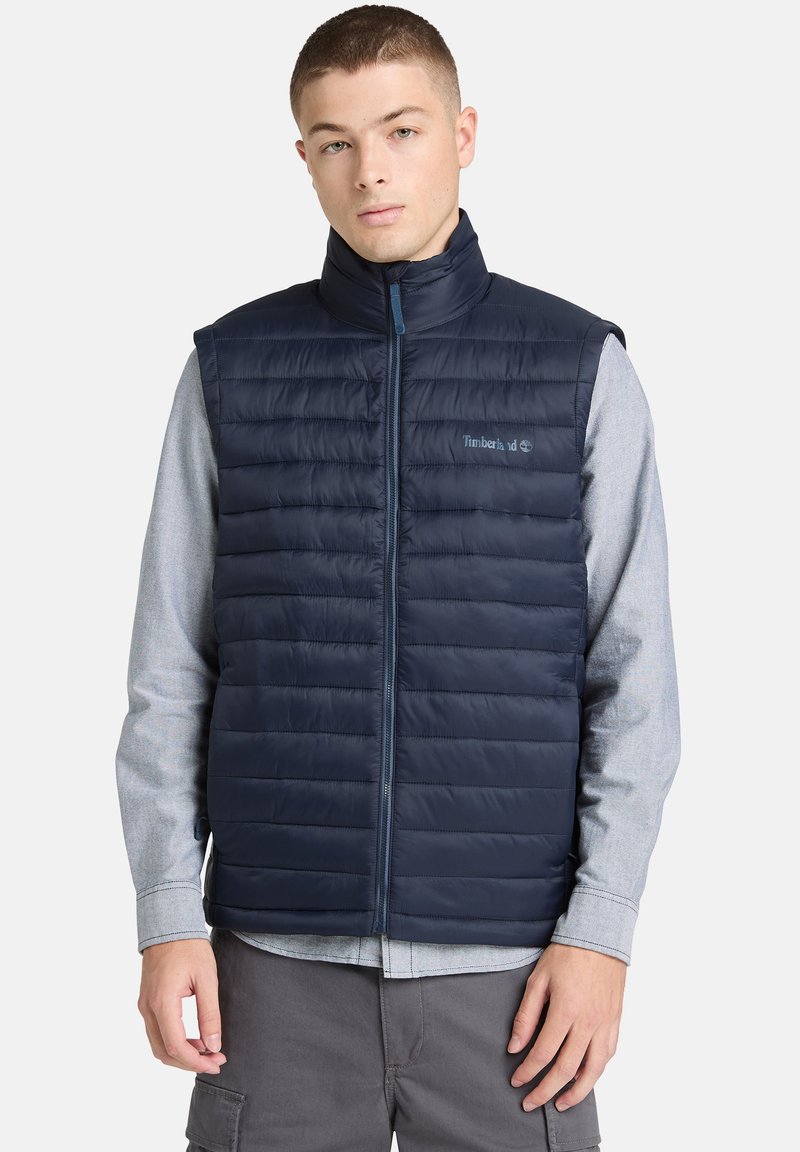 Giovane uomo che indossa un gilet trapuntato blu navy senza maniche sopra una maglia a maniche lunghe grigio chiaro, in piedi su uno sfondo semplice.