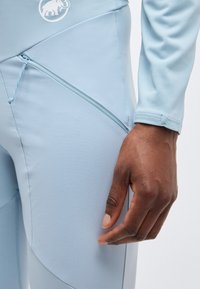 Tumša āda roka atrodas blakus gaiši ziliem sporta šortiem ar rāvējslēdzēja kabatu un baltu mamuta logotipu netālu no jostas.