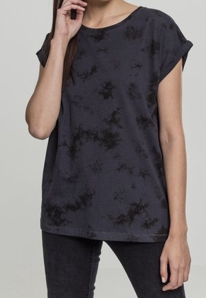 Kvinde iført en sort og mørkegrå tie-dye løs T-shirt med korte ærmer og op-rullede manchetter samt sorte tætsiddende bukser, hånd nær ansigtet.