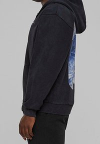 Sweat à capuche noir avec poignets et ourlet côtelés porté par une personne, présentant un graphisme circulaire bleu et blanc partiellement visible sur le dos.