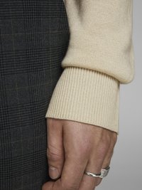 Manche de pull en tricot beige avec un poignet côtelé, à côté d'un tissu à carreaux sombres, mettant en valeur une main texturée avec une bague en argent.