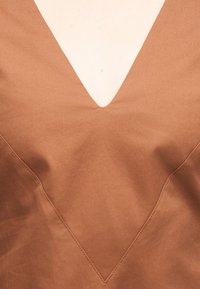 Καφέ ύφασμα με βαθύ V σχήματος ντεκολτέ που αποκαλύπτει ανοιχτόχρωμο δέρμα από κάτω, τονίζοντας τις λεπτομέρειες των ραφών κατά μήκος των άκρων του ντεκολτέ.