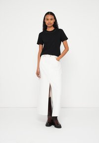 T-shirt nera a maniche corte con scollo borchiato, abbinata a una lunga gonna di jeans bianca con spacco frontale e stivali marroni alla caviglia.