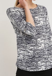 White blouse with navy circular pattern, round neckline, and three-quarter sleeves, featuring a smooth fabric and subtle shine.