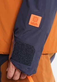 Manche de veste orange et marine avec un tissu texturé, un patch en velcro et un petit logo carré avec "ULLR" en relief.