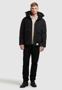 Czarna ocieplana kurtka z kapturem, z dwoma przednimi kieszeniami, zapięciem na zamek i sznurkiem ściągającym. Noszona na beżowym hoodie i czarnych spodniach.