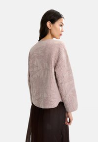 Kobieta z ciemnymi włosami, nosząca jasnoróżową, fakturowaną bluzkę z długim rękawem i przezroczystą, ciemną spódnicę, ustawiona w profilu na białym tle.