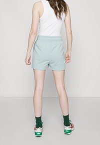 Woman wearing light blue shorts, white sleeveless top, green socks, and multicolored sneakers, standing facing away against a plain background.