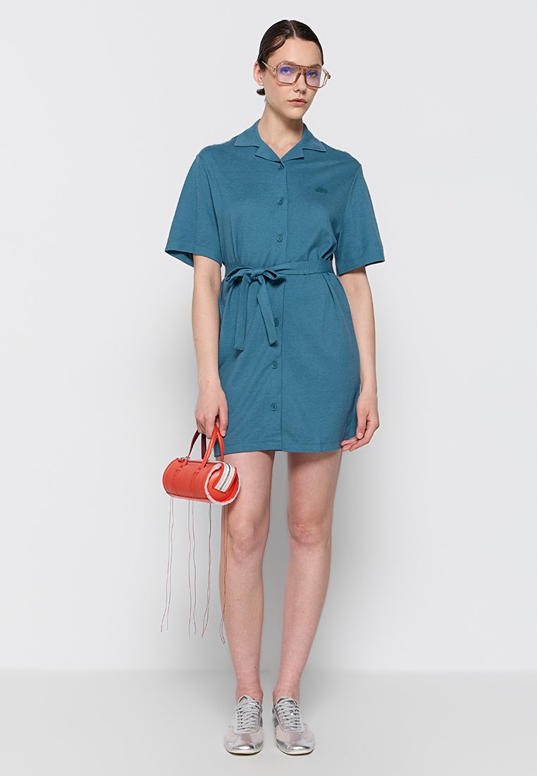Sukienka w kolorze turkusowym z krótkim rękawem, z zapięciem na guziki i paskiem w talii. Modelka trzyma małą pomarańczową torbę z białymi akcentami. Szare sneakersy dopełniają całość.