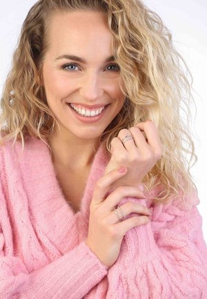 Krullend blond haar, lachende vrouw in een roze chunky gebreide trui, met meerdere zilveren ringen om haar vingers, tegen een witte achtergrond.