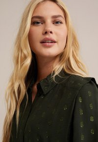 Camicetta button-up verde scuro con un tessuto testurizzato e motivi lucidi. Presenta un colletto standard e una vestibilità leggermente rilassata.
