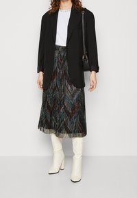 Černý blazer přes bílý tričko, doplněný o vícebarevnou plisovanou midi sukni s chevron vzorem a bílé vysoké boty.