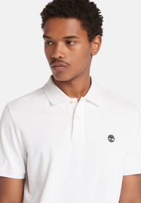 Bílá polo tričko vyrobené z bavlny. Má klasický límec, dvouknoflíkovou légu a malou námořnickou logotyp na levé straně hrudi. Krátké rukávy.