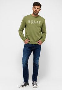Zelená bavlnená mikina s textom "MUSTANG", kombinovaná s modrými riflami a čiernymi teniskami s bielymi akcentmi, s voľným strihom.