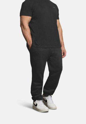 Homme portant un t-shirt noir, un pantalon de jogging noir et des baskets Nike blanches avec un swoosh noir, debout les mains partiellement dans les poches.
