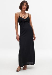 Schwarzes Maxikleid aus einem strukturierten, gerippten Stoff, mit Spaghettiträgern und einem Knoten-Detail an der Brust. Kombiniert mit silbernen Sandalen und geschichteter Schmuck.