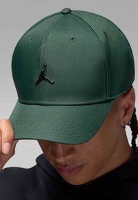 Ciemnozielona czapka wykonana z teksturowanej tkaniny, z czarnym logo Jumpman z przodu. Projekt zawiera zagięty daszek i guzik na górze.