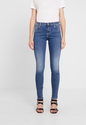 Woman wearing blue skinny jeans, white short-sleeve t-shirt, and black strappy heels standing against a plain background.