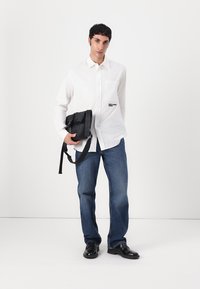 White button-up shirt with chest pocket, black crossbody bag, blue jeans, black shoes, model standing against a plain white background.