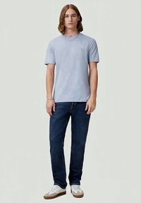 Νεαρός άνδρας που στέκεται φορώντας ένα ανοιχτό μπλε μπλουζάκι, σκούρα μπλε τζιν, λευκά αθλητικά παπούτσια, με καστανά μαλλιά μέχρι τους ώμους, μπροστά σε έναν απλό φόντο.