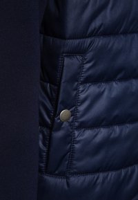 Marineblå quiltad jacka med ett slätt, glänsande tyg och en sidoficka som säkras med en silverfärgad tryckknapp, kontrasterad av en stickad del.