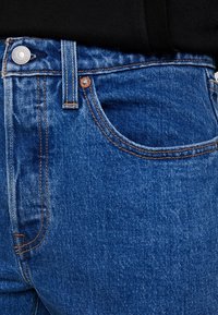 Jeans de mezclilla azul con un diseño clásico que incluye un cierre de botón y cremallera, un bolsillo frontal y detalles de costura en naranja contrastante.