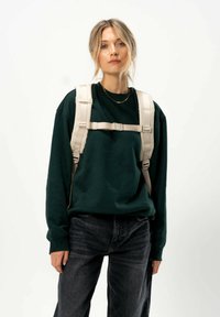 Un sac à dos harnais beige porté sur un sweat à capuche vert foncé, avec des bretelles épaisses et un matériel ajustable, associé à un jean noir.