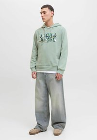 Mintgrøn hætteklædte sweatshirt med grafisk print, front kangaroo-lomme, parret med bredbenede, falmede denimjeans og beige slip-on sko.