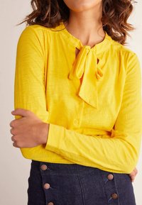 Blusa amarilla de manga larga con un lazo en el cuello, con botones en la parte frontal y hecha de una tela suave y texturizada, llevada sobre una falda de mezclilla oscura con botones metálicos.