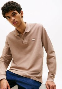 Beige Langarm-Polohemd aus strukturiertem Stoff, mit einer Zwei-Knopf-Leiste und einem kleinen Logo-Patch auf der Brust. Getragen mit blauen Jeans.