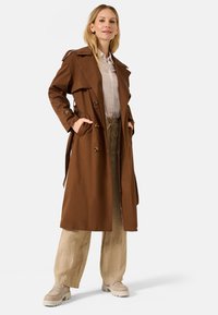 Cappotto trench marrone con vita con cintura, ampie revers e finiture dorate. Abbinato a pantaloni leggeri in lino e scarpe con plateau beige.