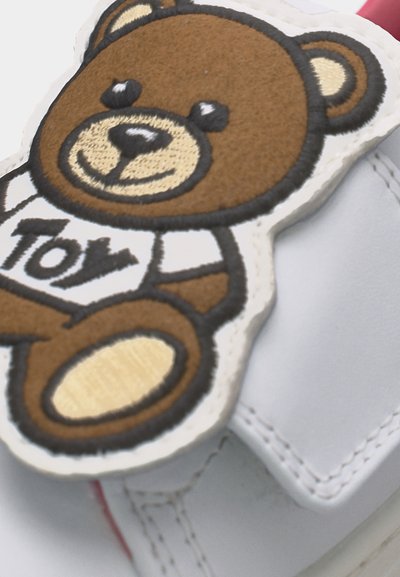 Κοντινή λήψη ενός λευκού αθλητικού παπουτσιού με ραμμένη καφέ φιγούρα αρκουδάκι, που φοράει πουκάμισο με τη λέξη "Toy".
