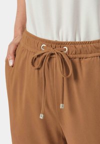 Pantalon en brun à cordon de serrage avec une texture douce, présentant une taille élastique avec des œillets en métal argenté et des bouchons.