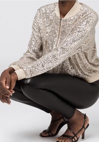 Pailletten Bomberjacke in Gold mit einem cremefarbenen, gerippten Kragen und Bündchen; verfügt über einen Reißverschluss vorne und eine geraffte Textur. Kombiniert mit glänzenden schwarzen Leggings.