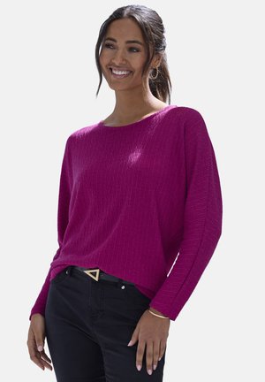 Donna sorridente con capelli scuri raccolti in una coda di cavallo, che indossa un maglione a costine magenta, pantaloni neri, fibbia della cintura a forma di triangolo e orecchini a cerchio dorati.