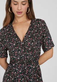 Schwarzes Blumenkleid mit kurzen Ärmeln, ausgestattet mit einem tiefen V-Ausschnitt und einem Bindegürtel in der Taille. Kleine rosa und weiße Blumen auf einem dunklen Hintergrund.