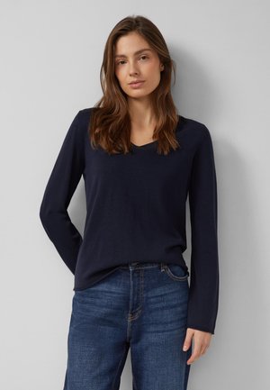 Frau mit langen braunen Haaren, die ein marineblaues langärmeliges Shirt und blaue Jeans trägt, steht vor einem hellgrauen Hintergrund.