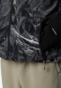 Svart och grå mönstrad jacka med en texturerad tyg, dragkedjestängning och en svart WindWall-accent. Bärs över en ljus beige fleece.