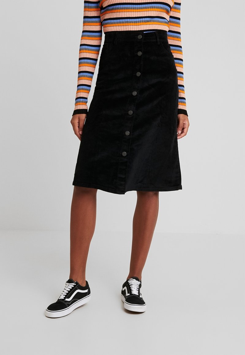 Femme portant une jupe noire boutonnée jusqu'aux genoux, un haut à manches longues rayé orange, bleu et noir, et des baskets noires et blanches.