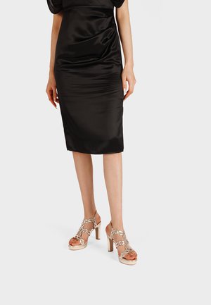 Donna che indossa una gonna a matita midi in satin nero e sandali beige con tacco alto e design con ritagli circolari, in piedi su uno sfondo bianco.