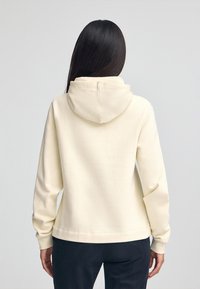 Krämfärgad hoodie med framficka, justerbar huva och ribbade muddar. Mjuk textur och avslappnad passform, kombinerad med mörka byxor.