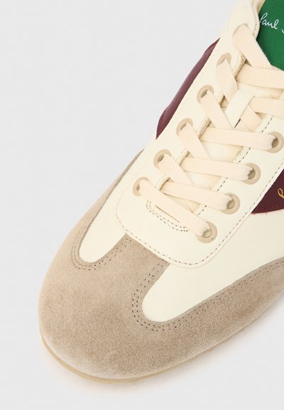 Κρεμ και μπεζ αθλητικό παπούτσι με κάλυμμα δέρματος σουέντ στη μύτη, μπεζ κορδόνια κ�αι μπορντό λεπτομέρειες. Η γλώσσα είναι πράσινη με αντίθετο κείμενο.